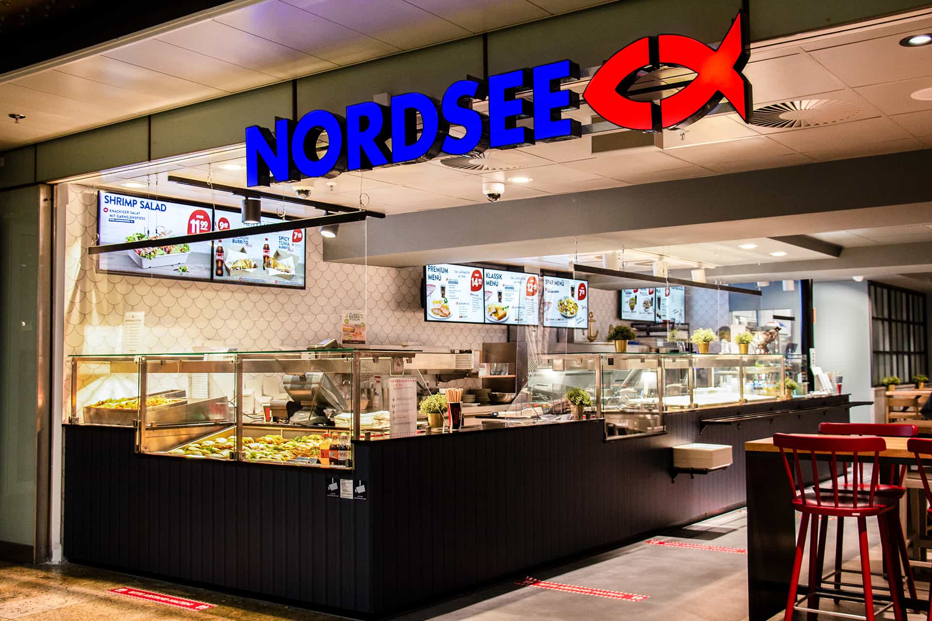 Nordsee Fischrestaurant mit beleuchtetem blauen Schriftzug und rotem Fischlogo, mit Essensausgabe und digitaler Speisekarte.