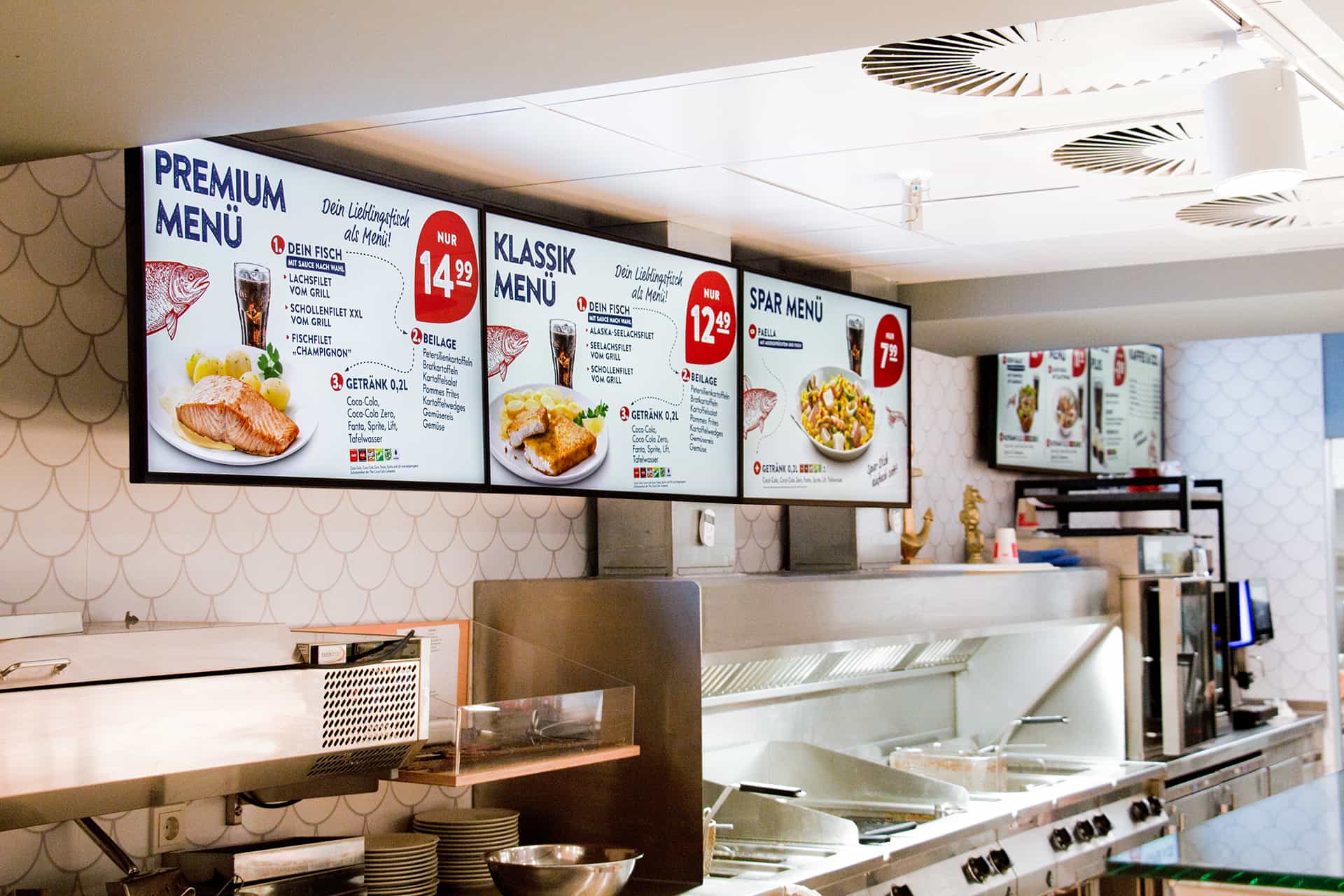Digitales Speisekarten-Display in einem Restaurant mit Premium-, Klassik- und Spar-Menüs, die Fischgerichte mit Beilagen und Getränken zeigen.