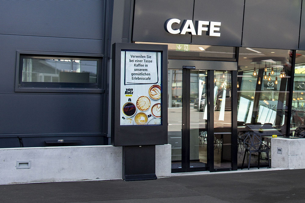 Außenbereich eines modernen Cafés mit digitalem Anzeigebildschirm, der eine Werbung für Kaffee mit dem Text 'Verweilen Sie bei einer Tasse Kaffee in unserem gemütlichen Erlebniscafé' zeigt.
