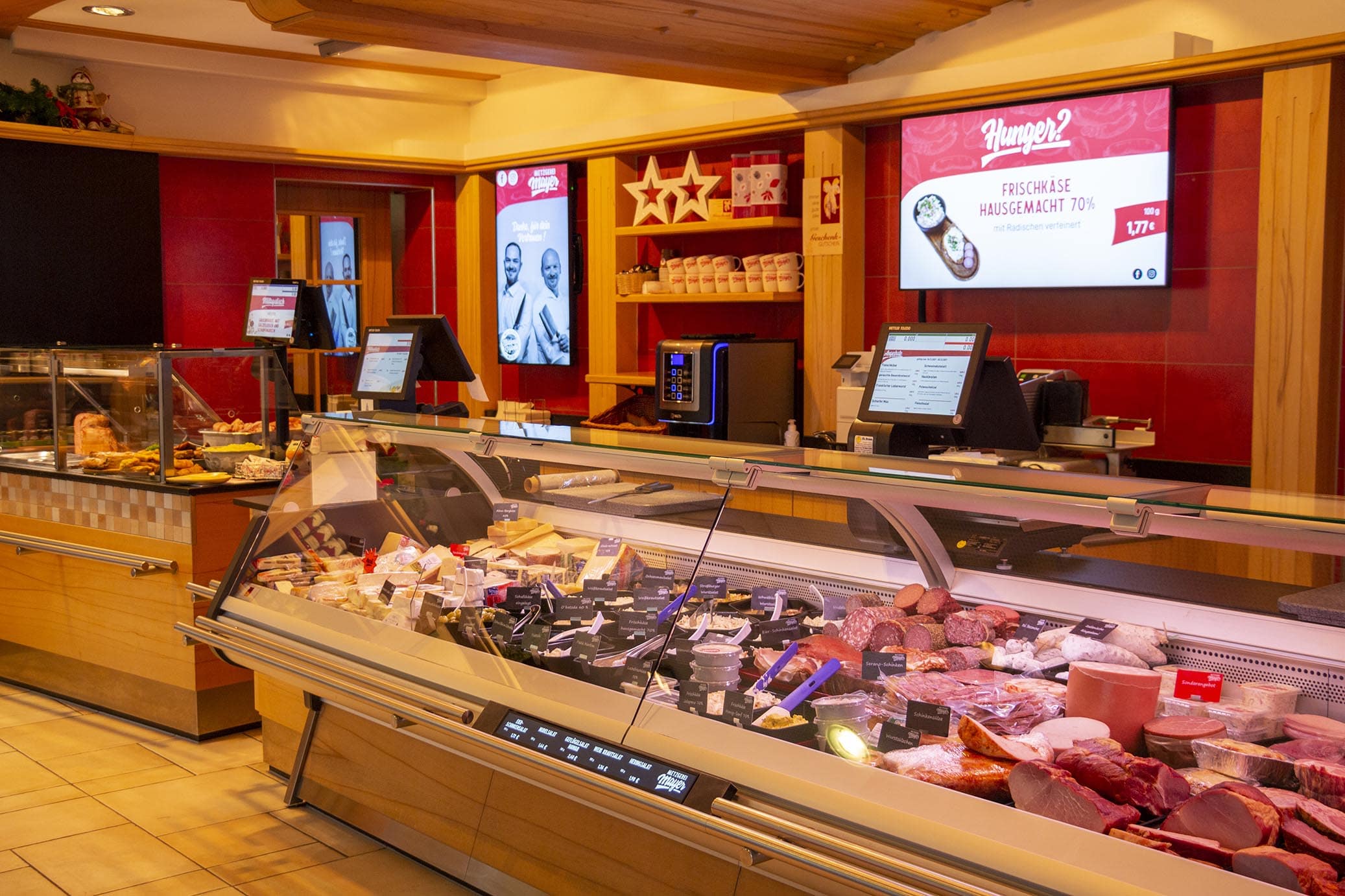 Moderne Metzgereitheke mit frischen Fleisch- und Wurstwaren, digitalen Bildschirmen und einer warmen Holzverkleidung.