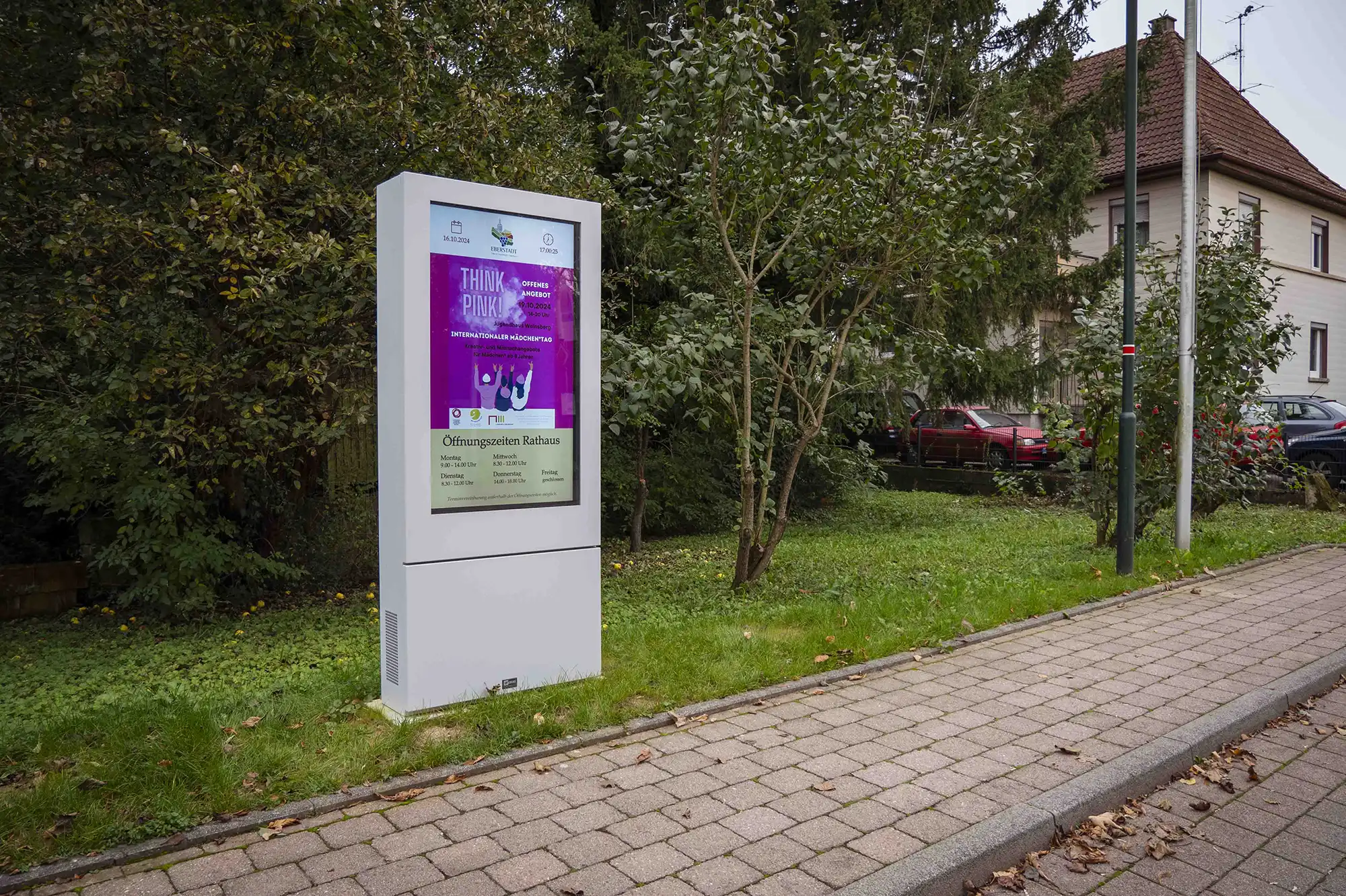 Digitale Informationsstele am Straßenrand mit einem pinkfarbenen Plakat und Informationen zu den Öffnungszeiten des Rathauses.
