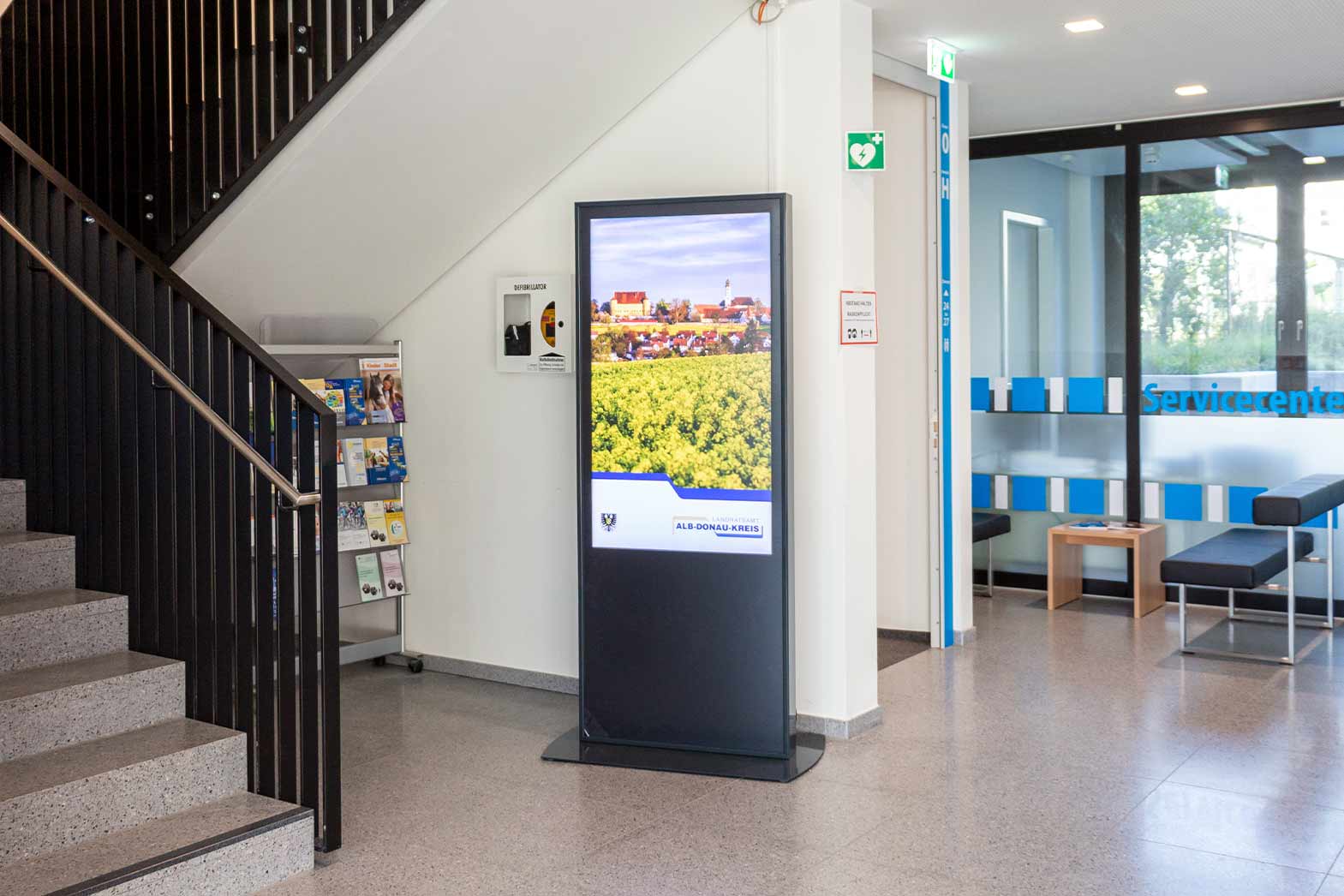 Empfangsbereich mit Treppe, Infoständer, einem digitalen Informationsbildschirm mit Landschaftsbild und Sitzgelegenheiten vor einem Glasabtrennung mit Schriftzug 'Servicecenter'.