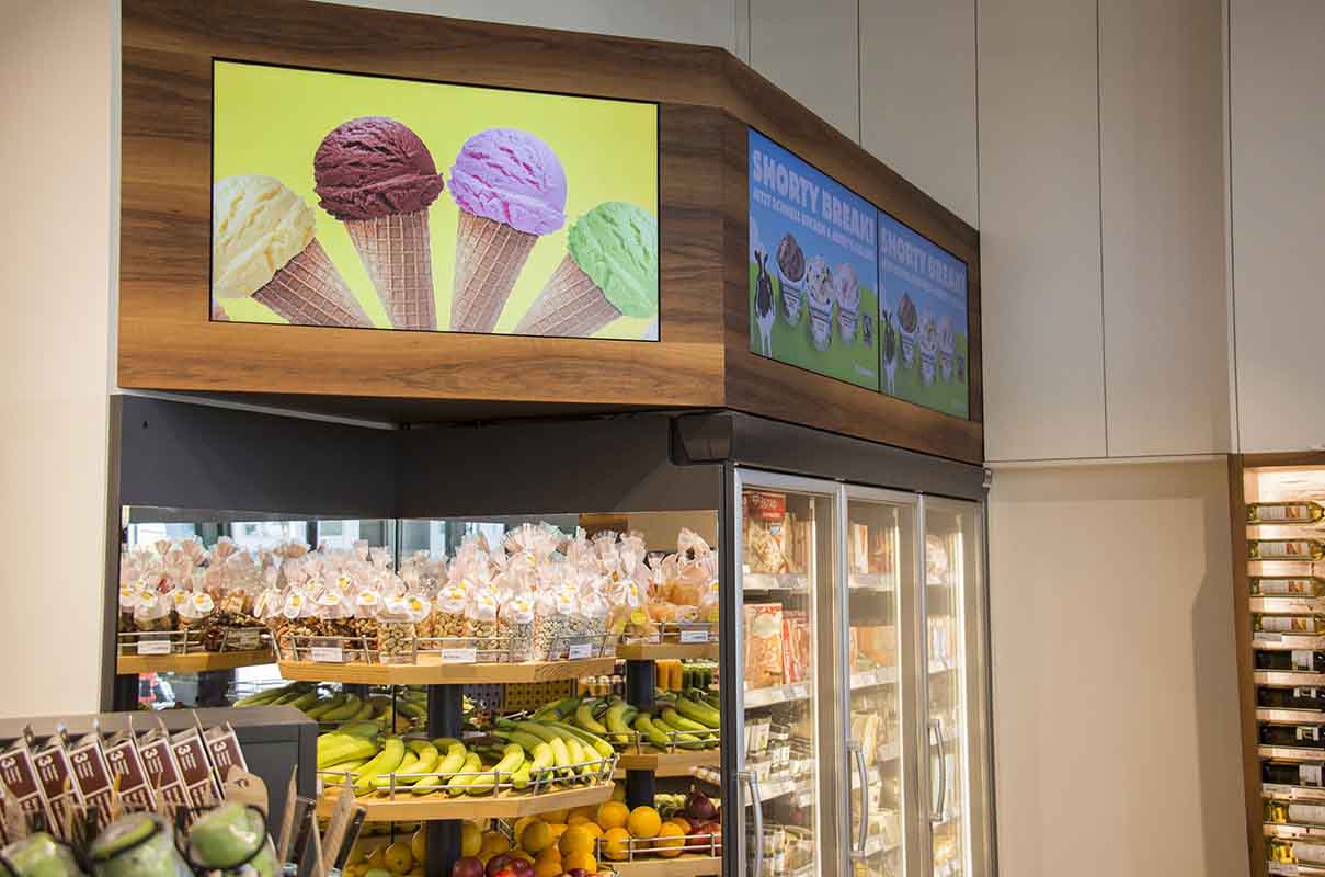 Kühlschrank in einem Geschäft mit Bananen, Orangen und verpackten Snacks, beleuchtet von digitalen Displays mit Eiscremebildern.