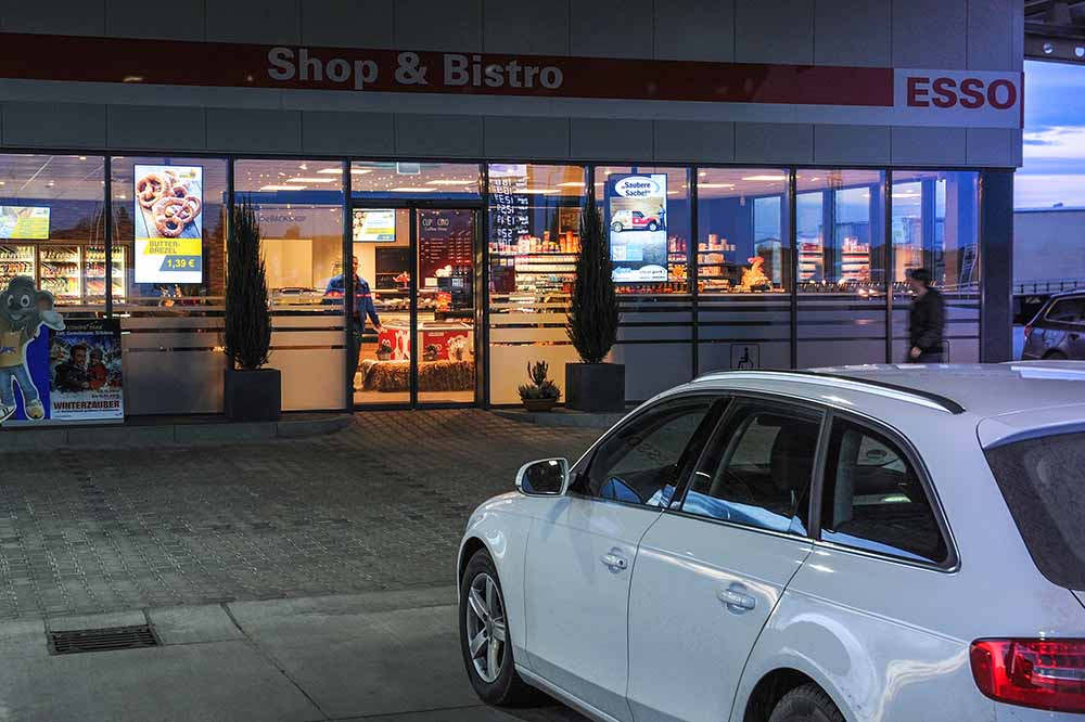 Außenansicht einer Esso-Tankstelle mit beleuchtetem Shop & Bistro, weißen parkenden Auto und Menschen im Hintergrund.
