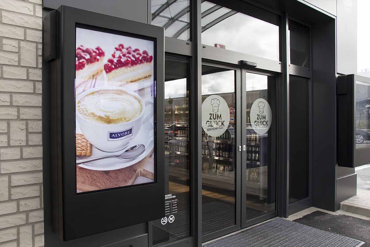 Digitale Anzeigetafel neben dem Eingang eines Geschäfts mit einem Bild von Kaffee, Gebäck und Kuchenstücken.