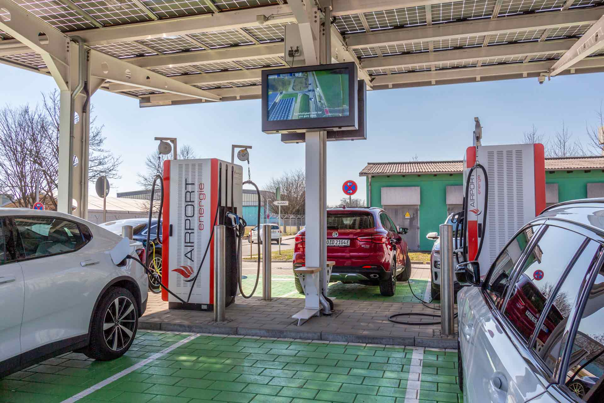 Elektroautos an zwei Airport Energie Ladestationen unter einem Dach mit Solarpanelen, ein Bildschirm ist darüber montiert.