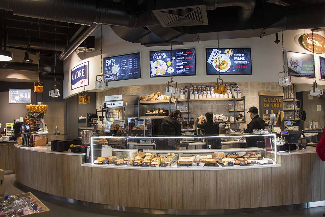 Moderne Bäckerei mit beleuchtetem Verkaufstresen voller Backwaren und Kaffee-Menüs auf digitalen Bildschirmen im Hintergrund.