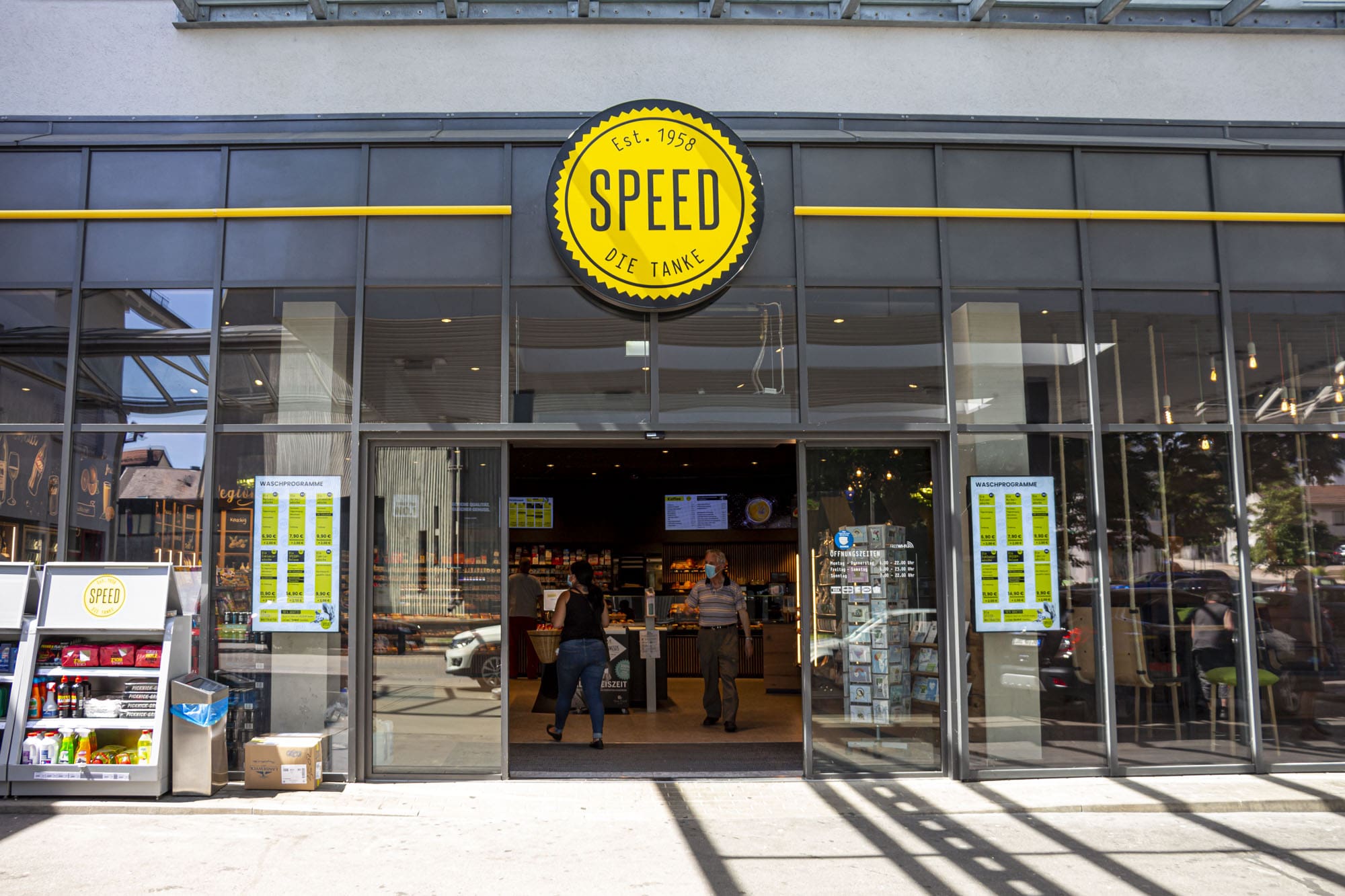 Außenansicht eines Geschäfts mit großer Glasfassade, Schild mit gelbem Logo 'SPEED DIE TANKE' und Menschen am Eingang.