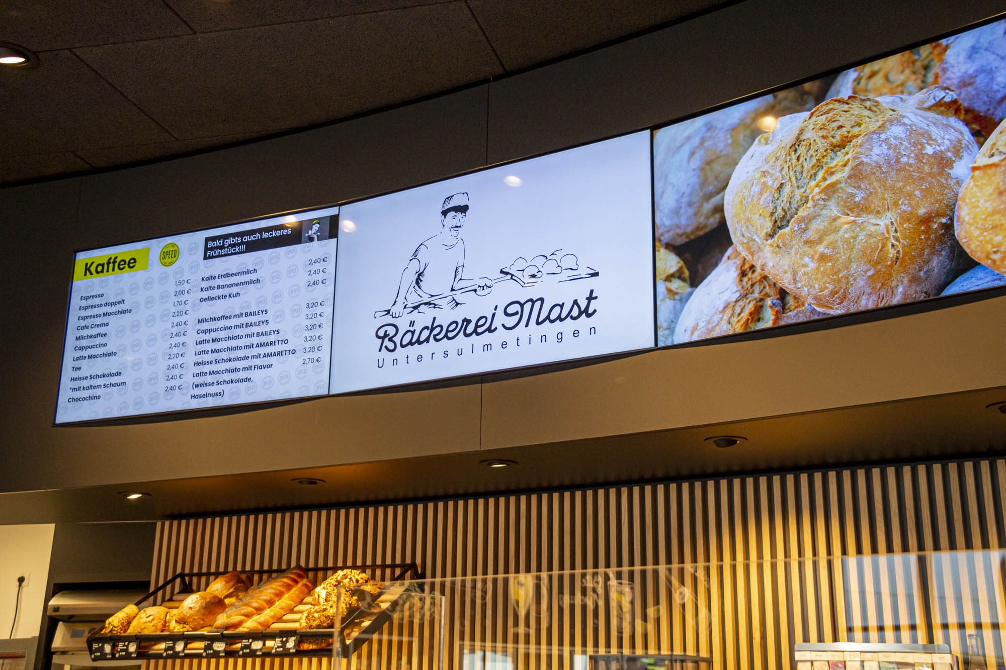 Digitales Menüboard einer Bäckerei mit Kaffeepreisübersicht, Bäckerei Mast Logo und Bild von frischem Brot.