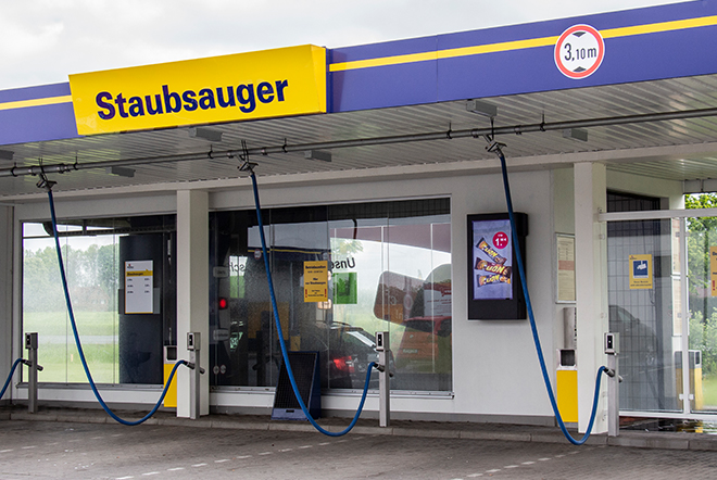 Selbstbedienungsstation mit drei blauen Staubsaugerschläuchen unter einem Dach mit dem gelben Schild ‚Staubsauger‘ an einer Tankstelle.