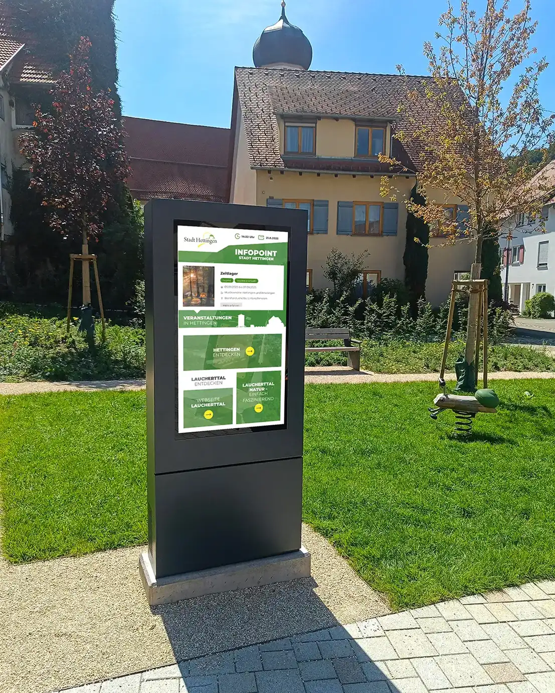 Digitale Infotafel im Freien mit Informationen zur Stadt Hettingen, umgeben von Grünflächen und traditionellen Gebäuden im Hintergrund.