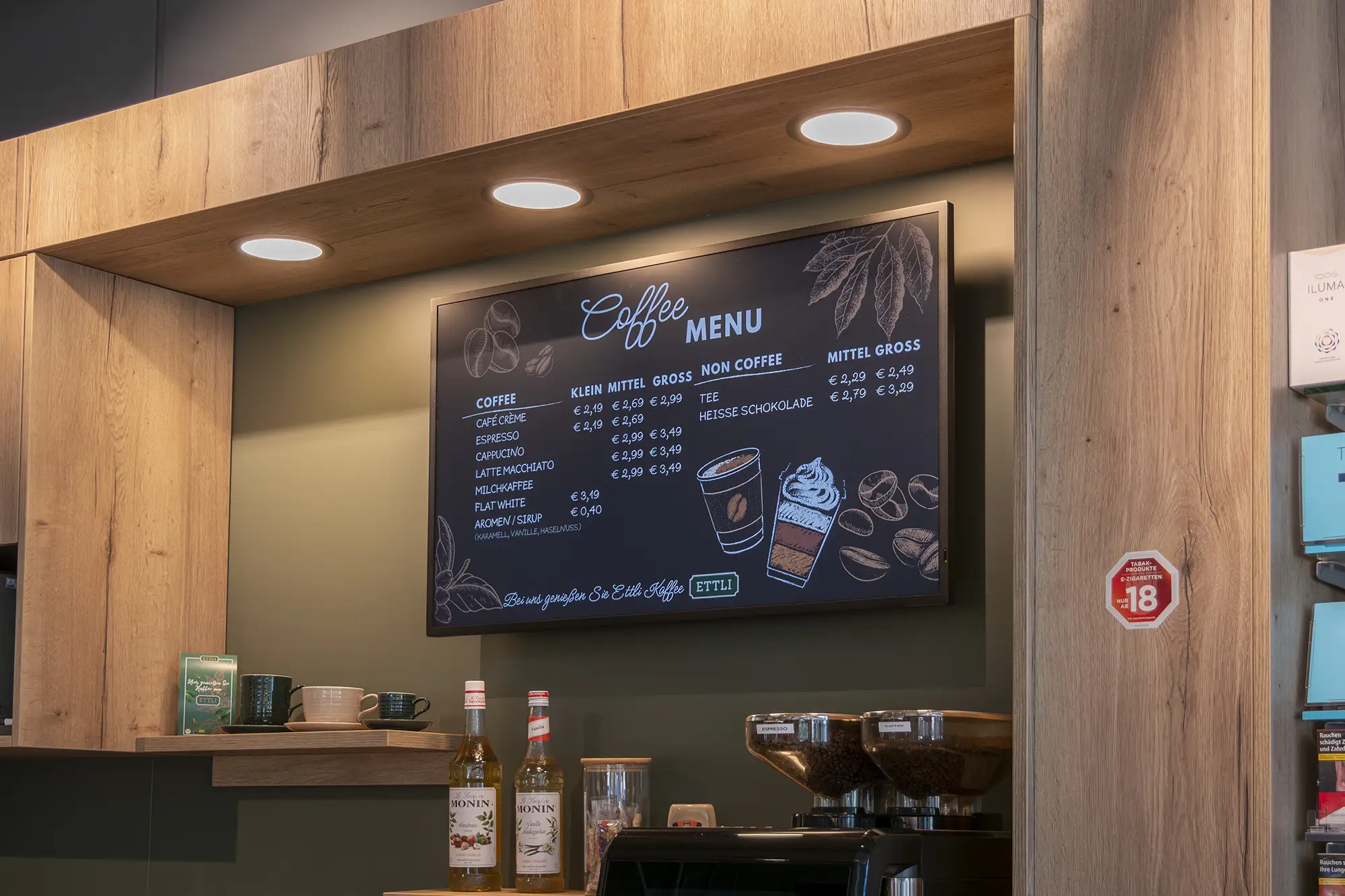 Digitales Coffee-Menü in einer Kaffeebar mit Preisen für verschiedene Kaffeesorten und Sirupflaschen auf der Theke.