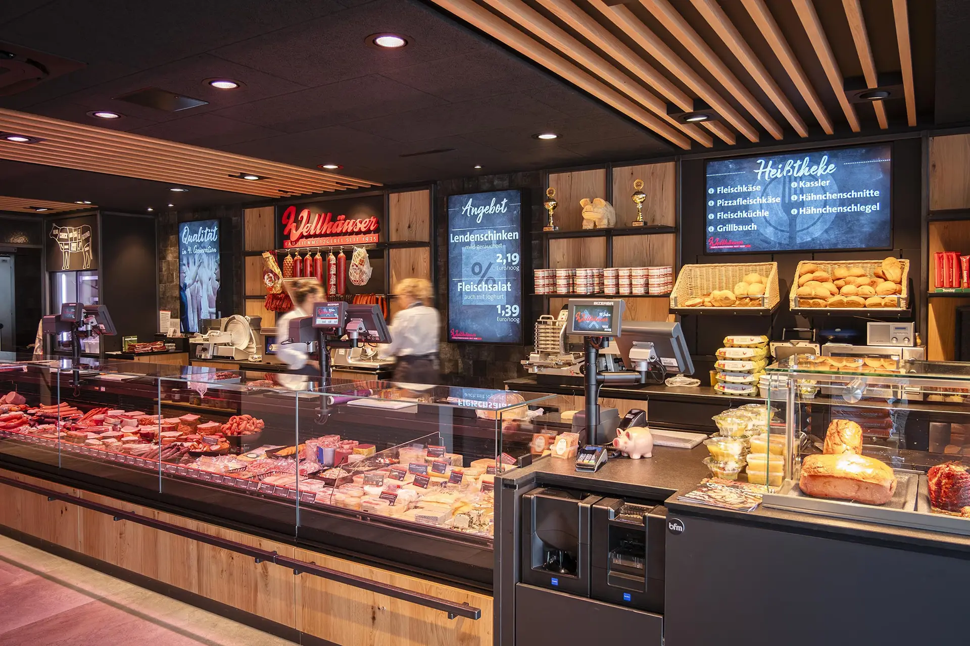 Moderne Metzgerei mit beleuchtetem Thekenbereich, verschiedenen Fleisch- und Wurstwaren, zwei Mitarbeiterinnen hinter der Theke und digitalen Preistafeln an der Wand.