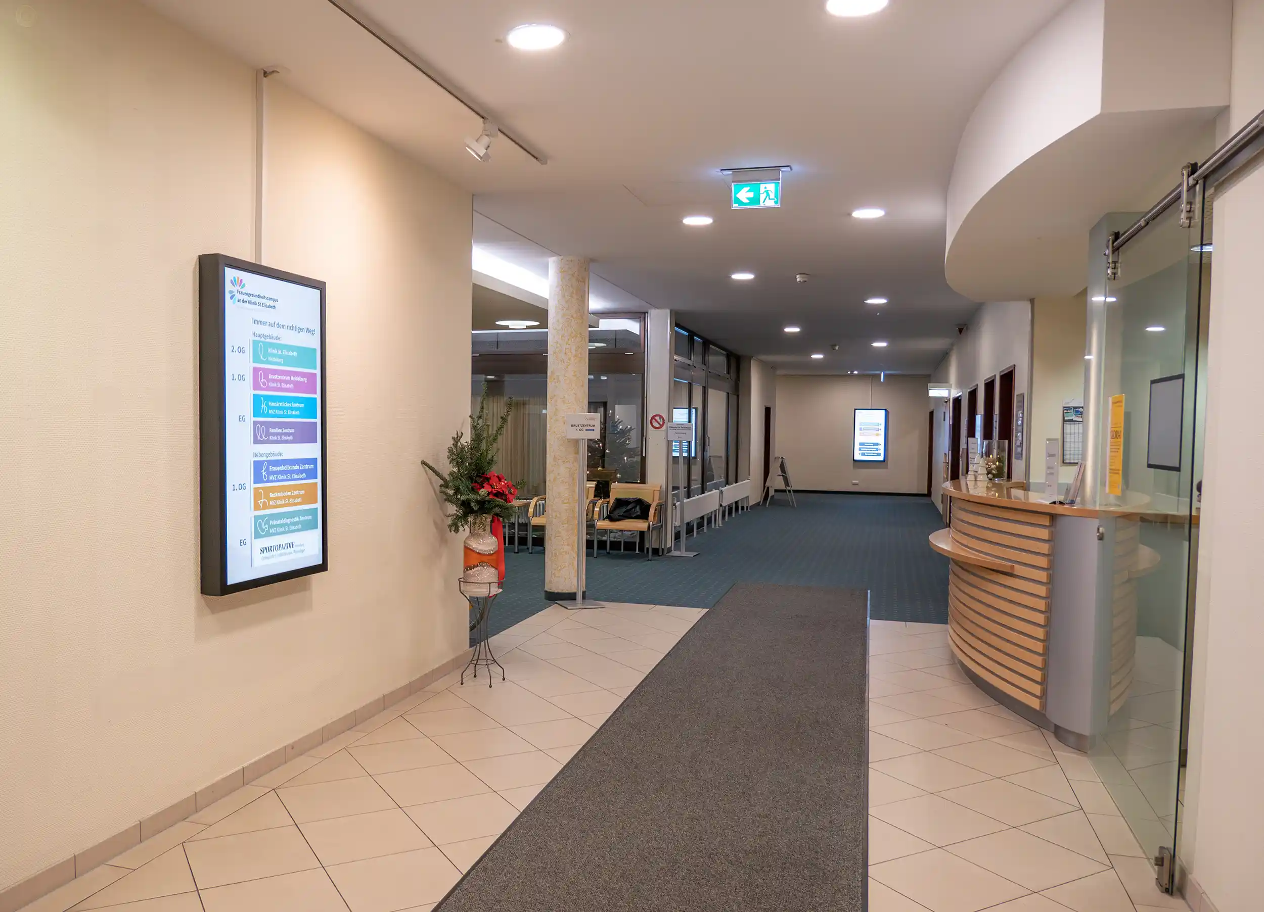 Innenflur eines medizinischen Gebäudes mit digitalem Verzeichnis an der Wand, Empfangstresen rechts und Sitzbereich im Hintergrund.