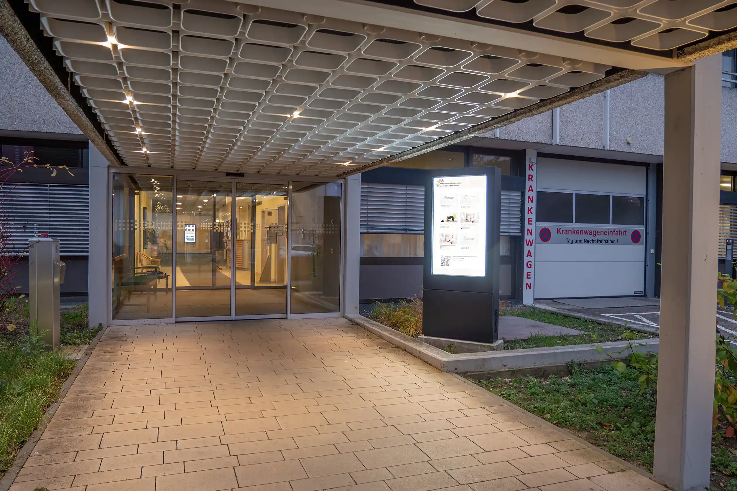 Eingang eines Krankenhauses mit automatischen Glastüren, beleuchtetem Informationsdisplay und Einfahrt für Krankenwagen.