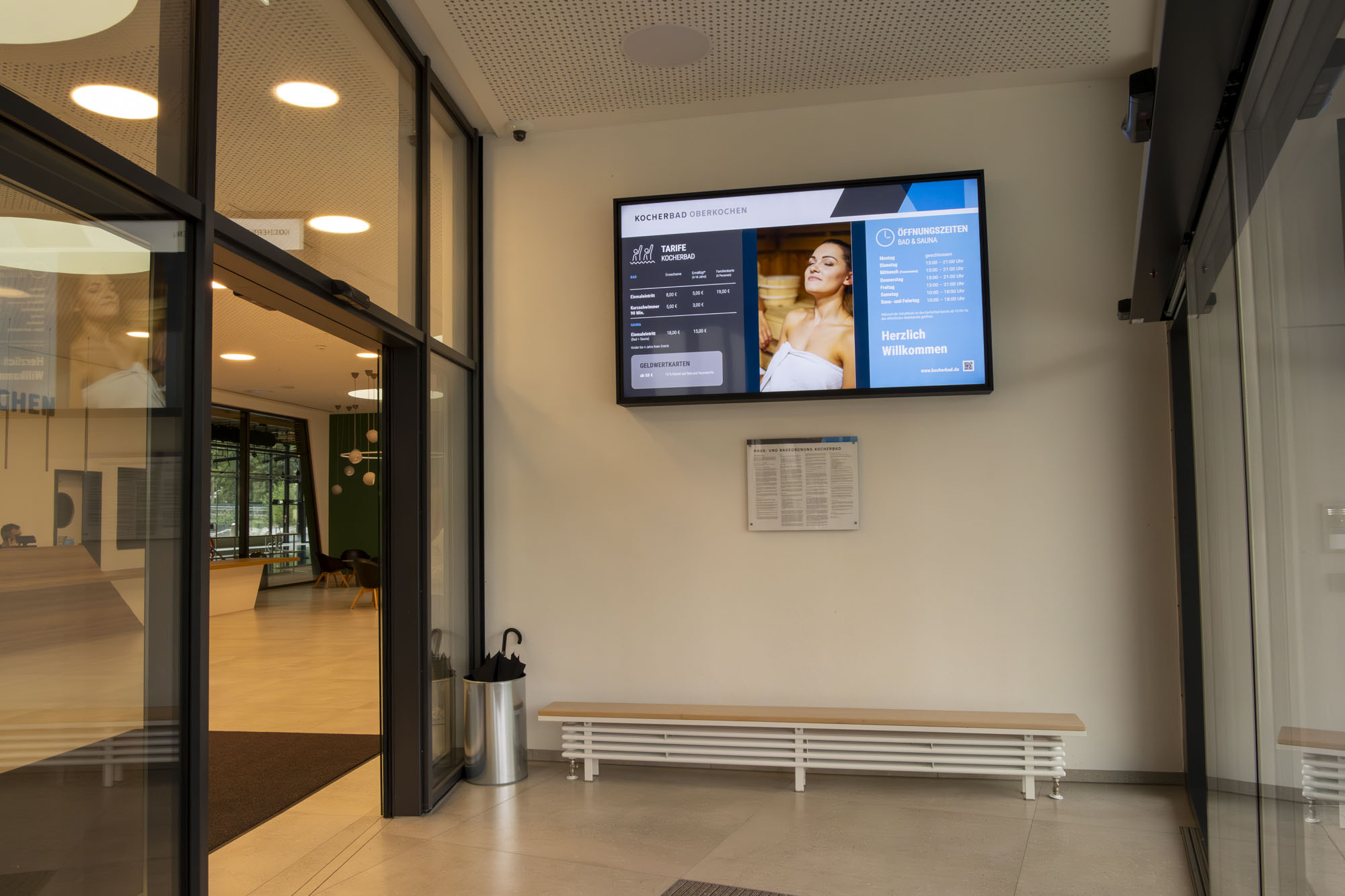 Eingangsbereich eines Gebäudes mit Sitzbank, Regenschirmständer und digitalem Informationsbildschirm mit Öffnungszeiten und Preisinformationen für das Kocherbad Oberkochen.