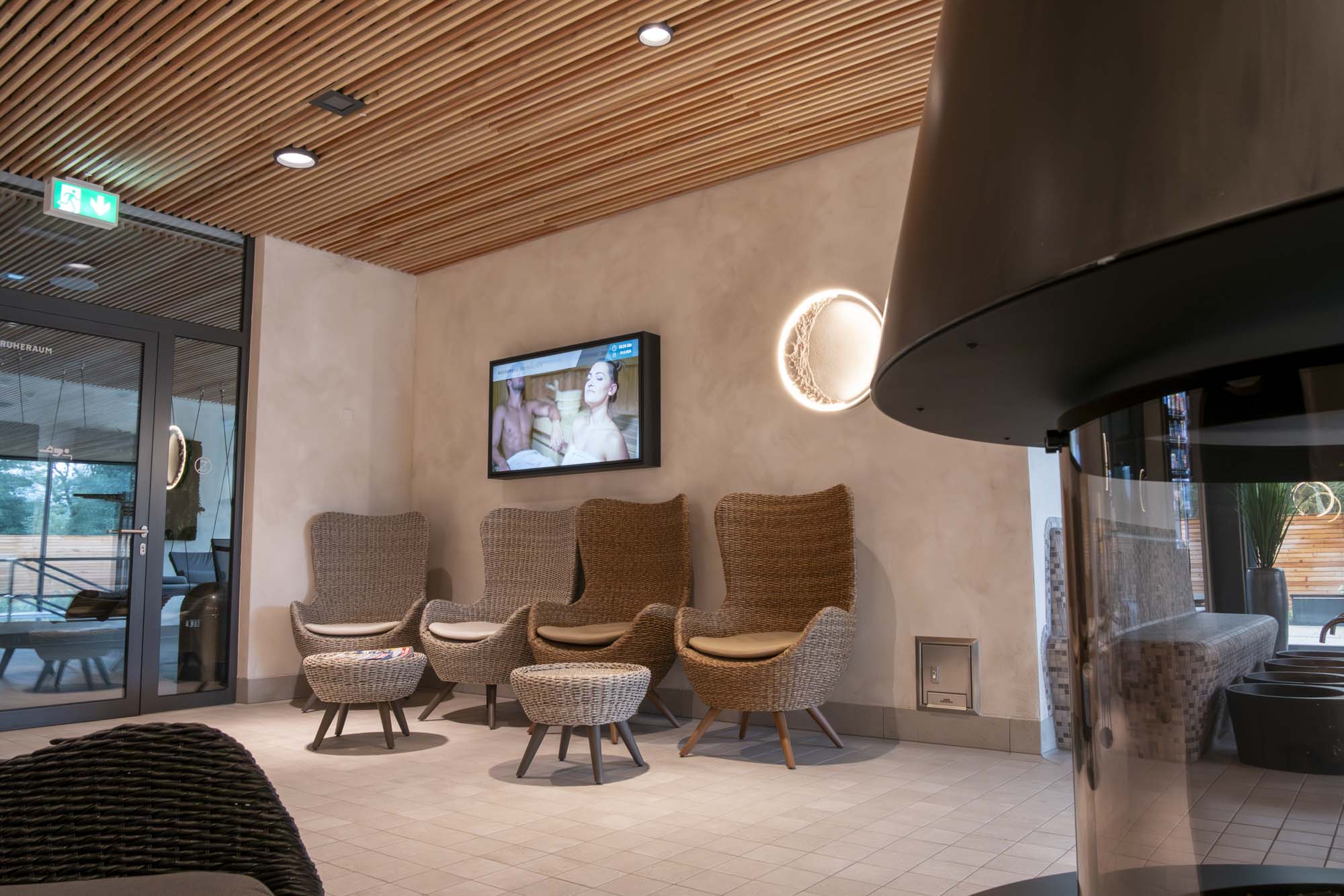 Moderner Wartebereich mit geflochtenen Sesseln und Poufs, digitalem Bildschirm an der Wand und rundem Wandlicht.