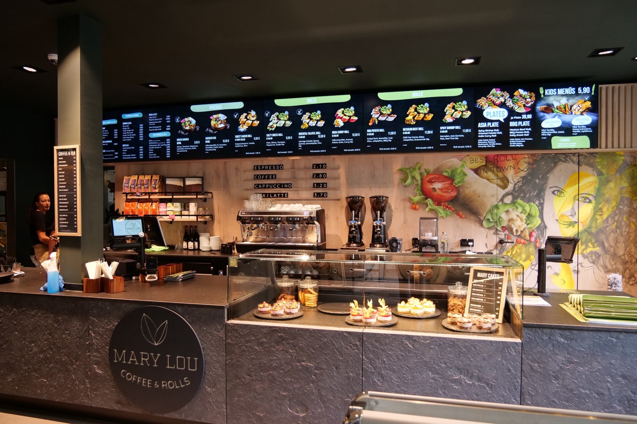 Innenansicht eines modernen Cafés mit Laptoppult, digitaler Speisekarte über der Theke und einem Kuchen- und Gebäckdisplay.