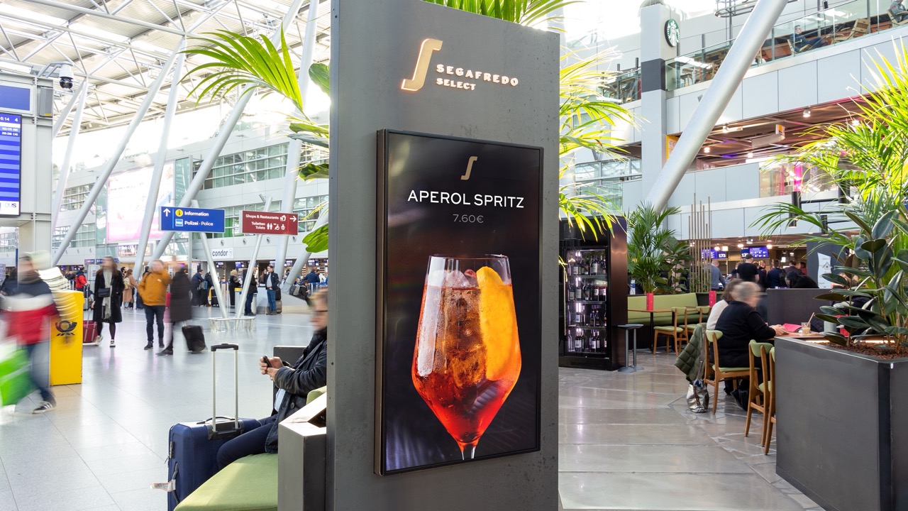 Werbeschild für Aperol Spritz am Flughafen mit Passagieren und Sitzbereich im Hintergrund.