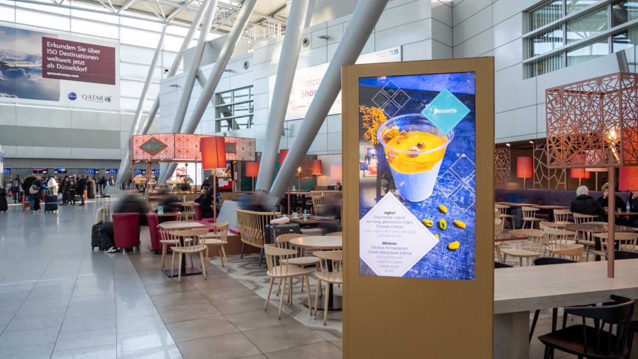 Digitales Werbedisplay zeigt ein Dessert mit Joghurt und Mango in einem modernen Flughafen-Restaurant mit Gästen und Sitzbereichen.
