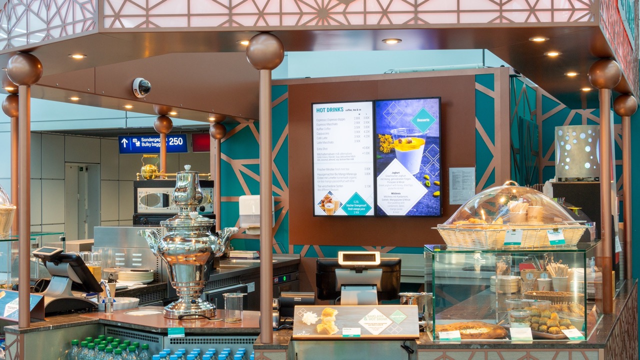 Kaffee- und Snackstand in einem Flughafen mit digitaler Menüanzeige und ausgestellten Backwaren hinter Glas.