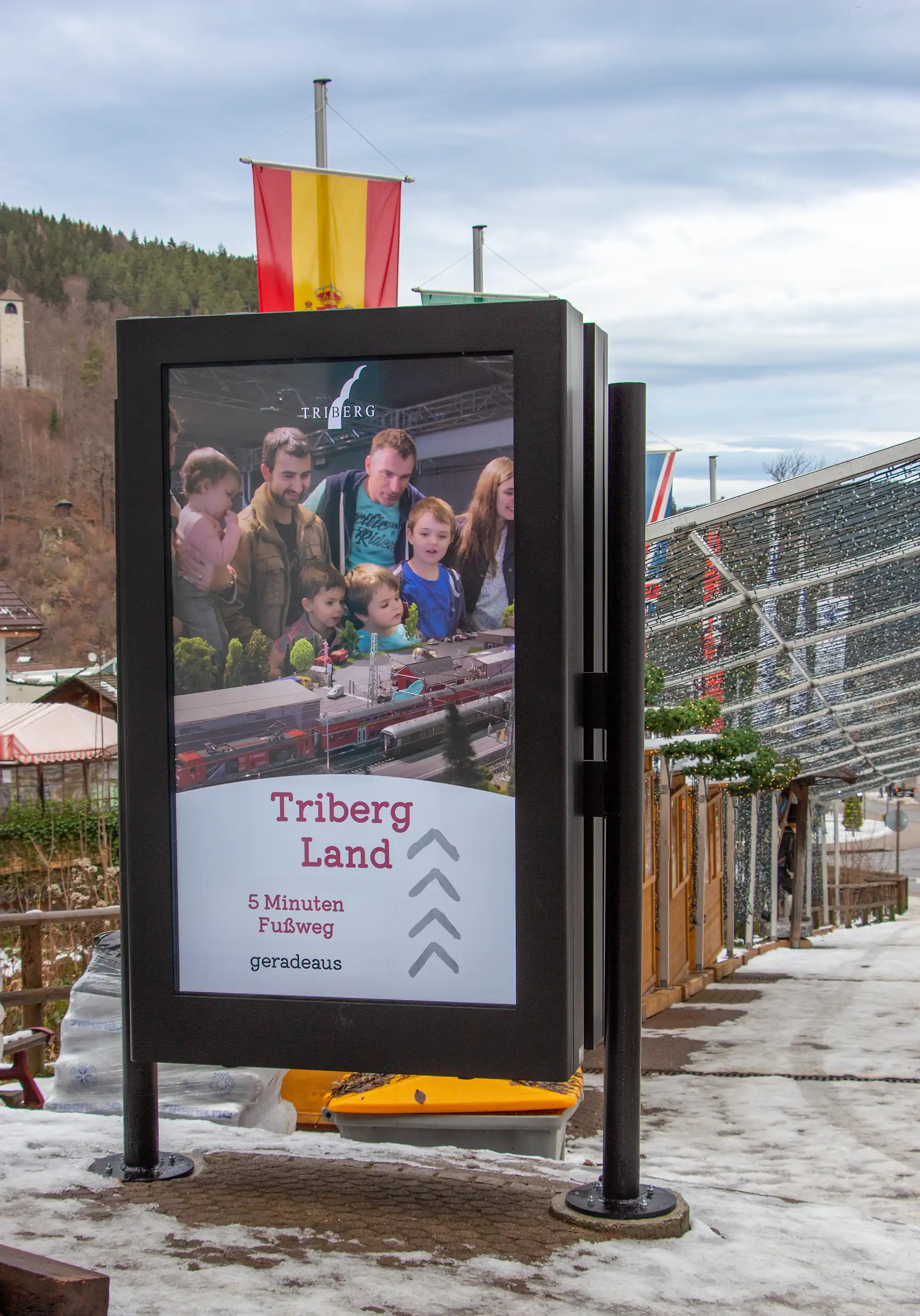 Digitale Informationstafel am Weg mit Werbung für Triberg Land, zeigt eine Gruppe von Kindern und Erwachsenen, die eine Modelleisenbahn betrachten.