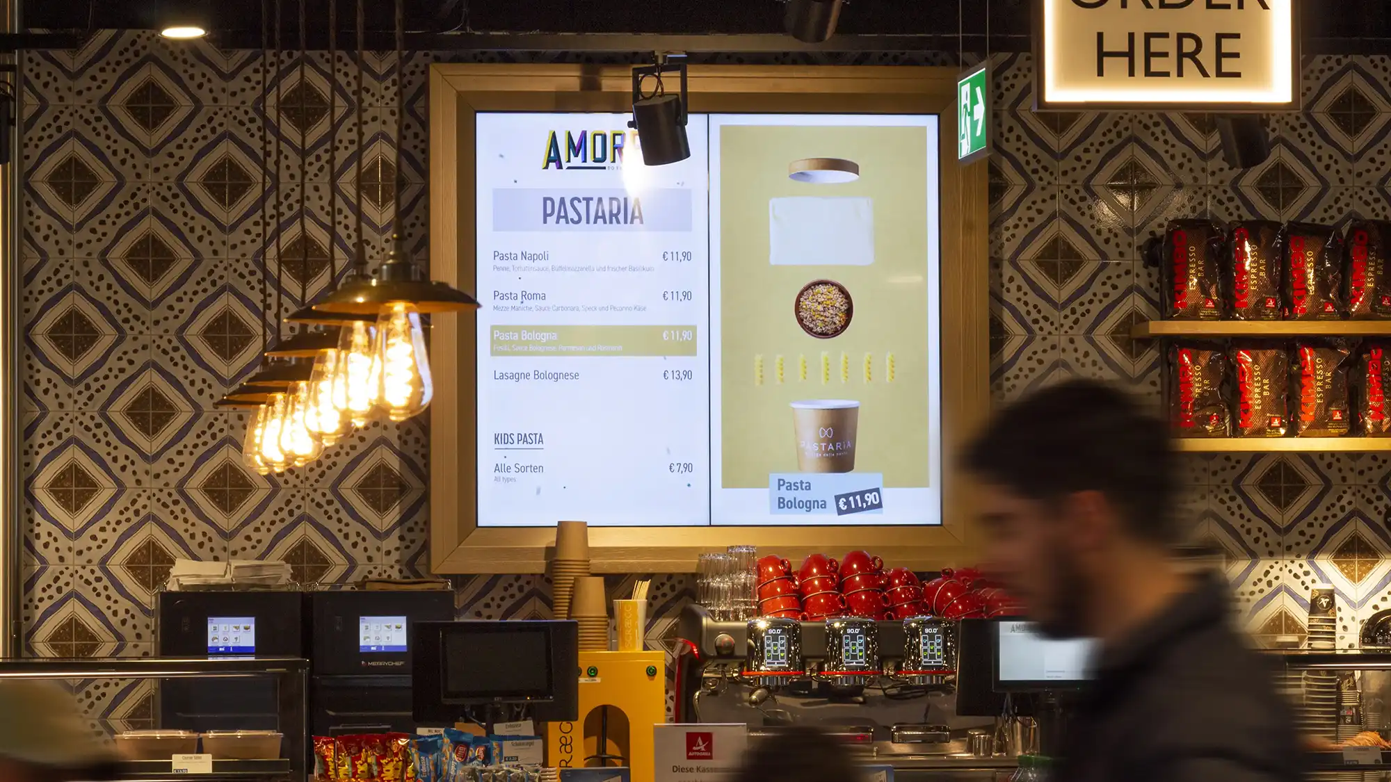 Digitales Menü in einem Restaurant mit Pasta-Gerichten und Preisanzeige, beleuchtete Retro-Lampen und verschwommener Mann im Vordergrund.