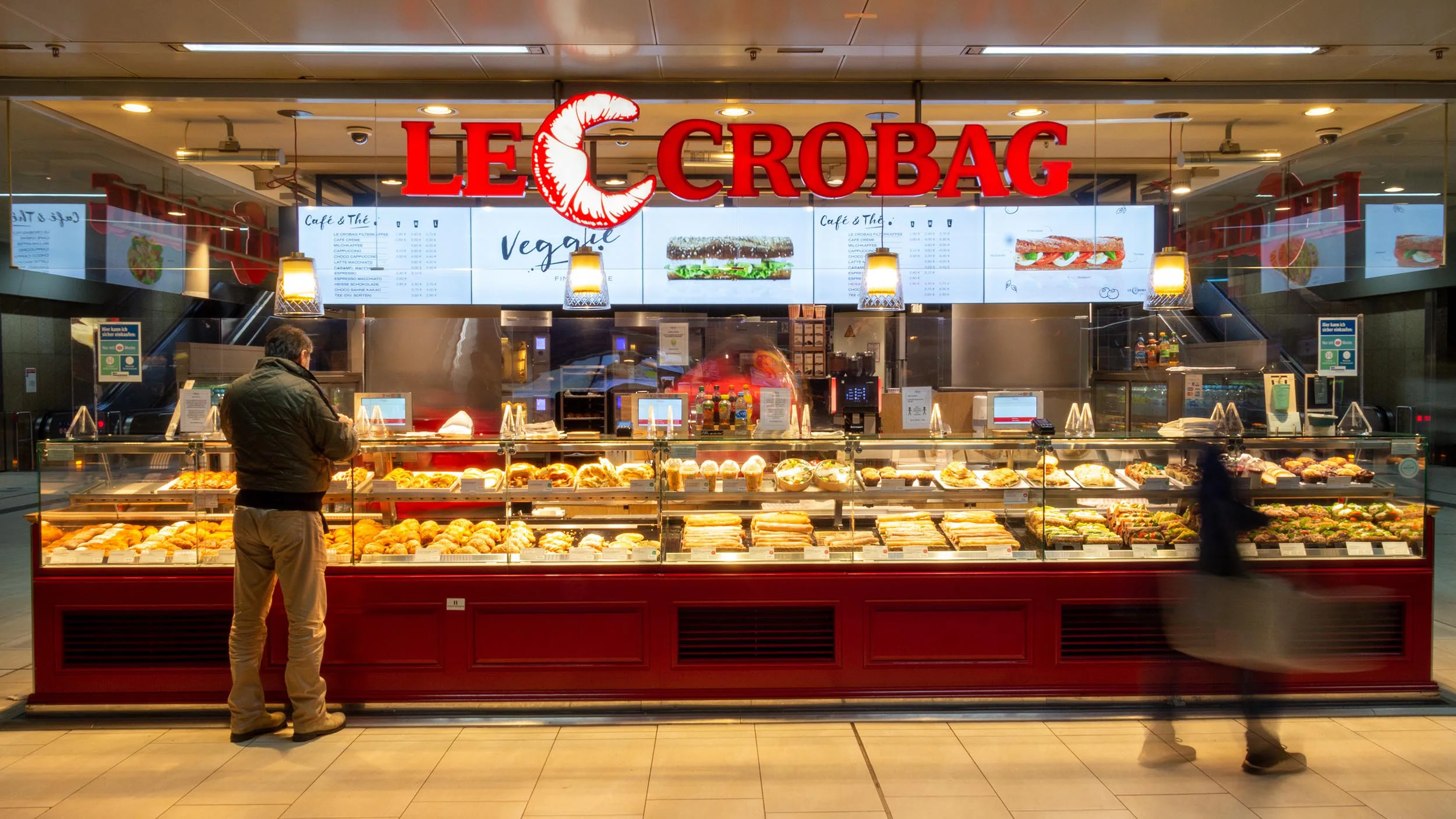 Bäckerei Le Crobag mit beleuchtetem roten Logo und einer Theke voller verschiedener Backwaren, davor ein Kunde und eine vorbeigehende Person.
