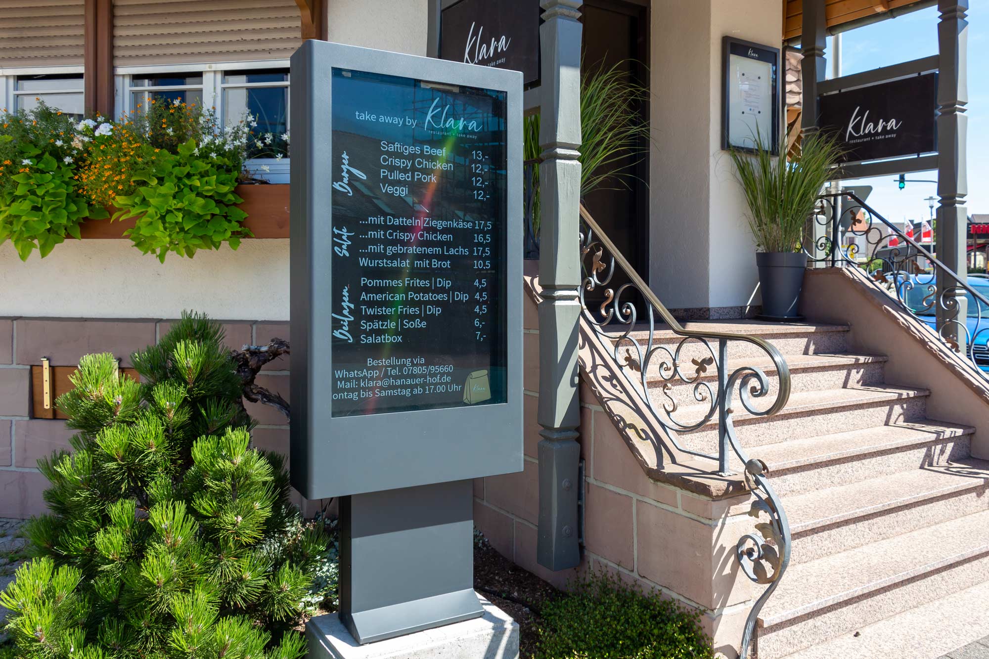 Außenbereich eines Restaurants mit digitalem Menüdisplay neben einer Treppe und blühenden Pflanzen.