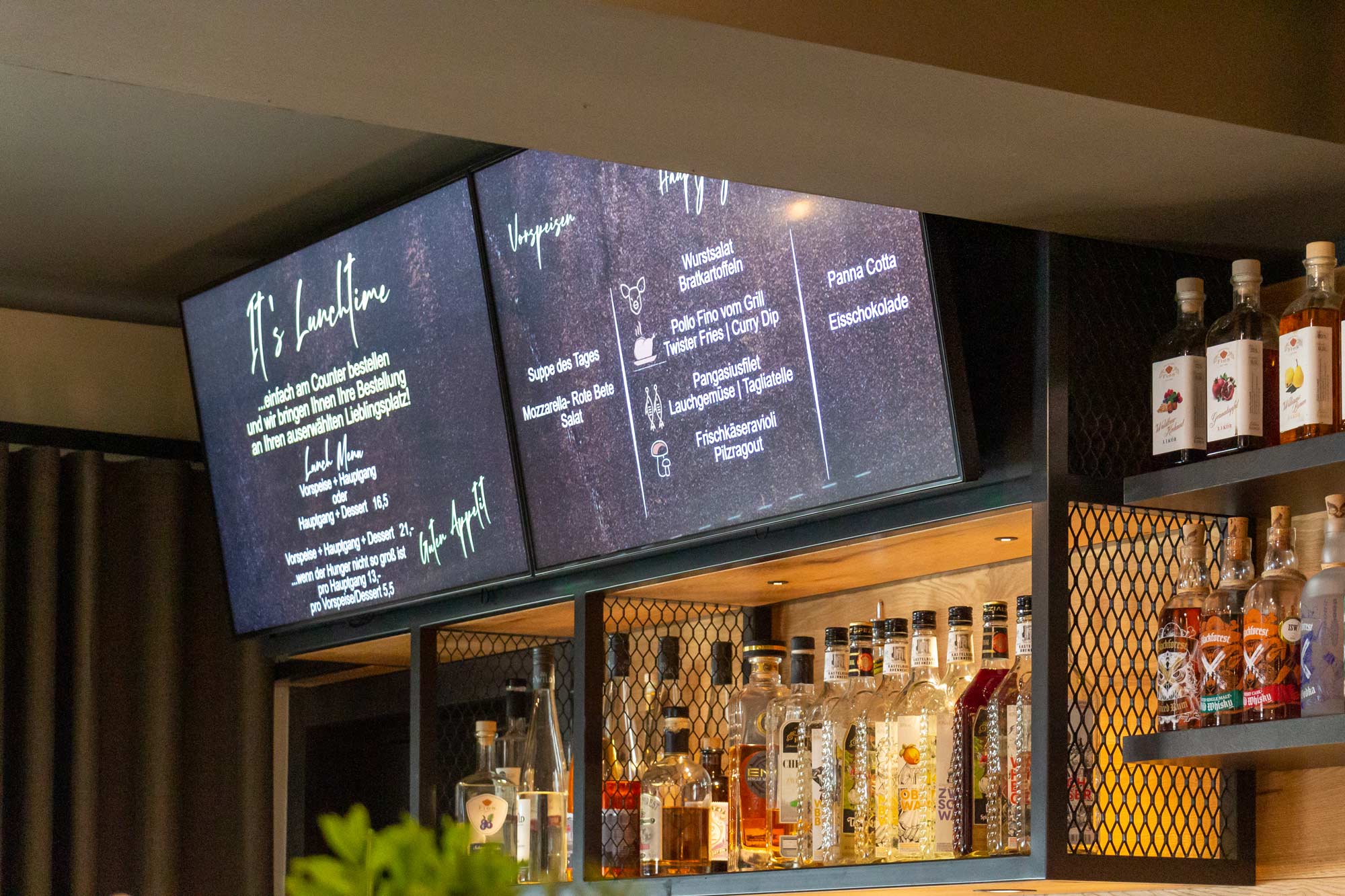 Digitales Menüdisplay über einer Bar mit Spirituosenflaschen, zeigt Mittagsmenü mit Vorspeisen, Hauptgerichten und Desserts in deutscher Sprache.