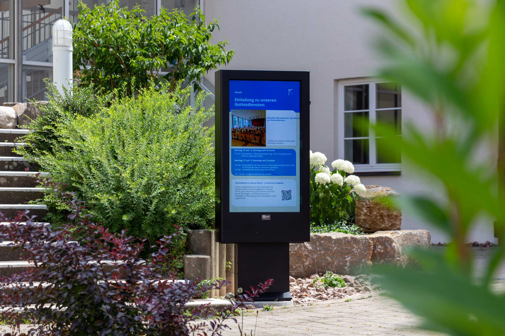 Digitales Informationsdisplay vor einem Gebäude zeigt Einladung zu Gottesdiensten mit Details und QR-Code, umgeben von grünen Pflanzen und weißen Blumen.