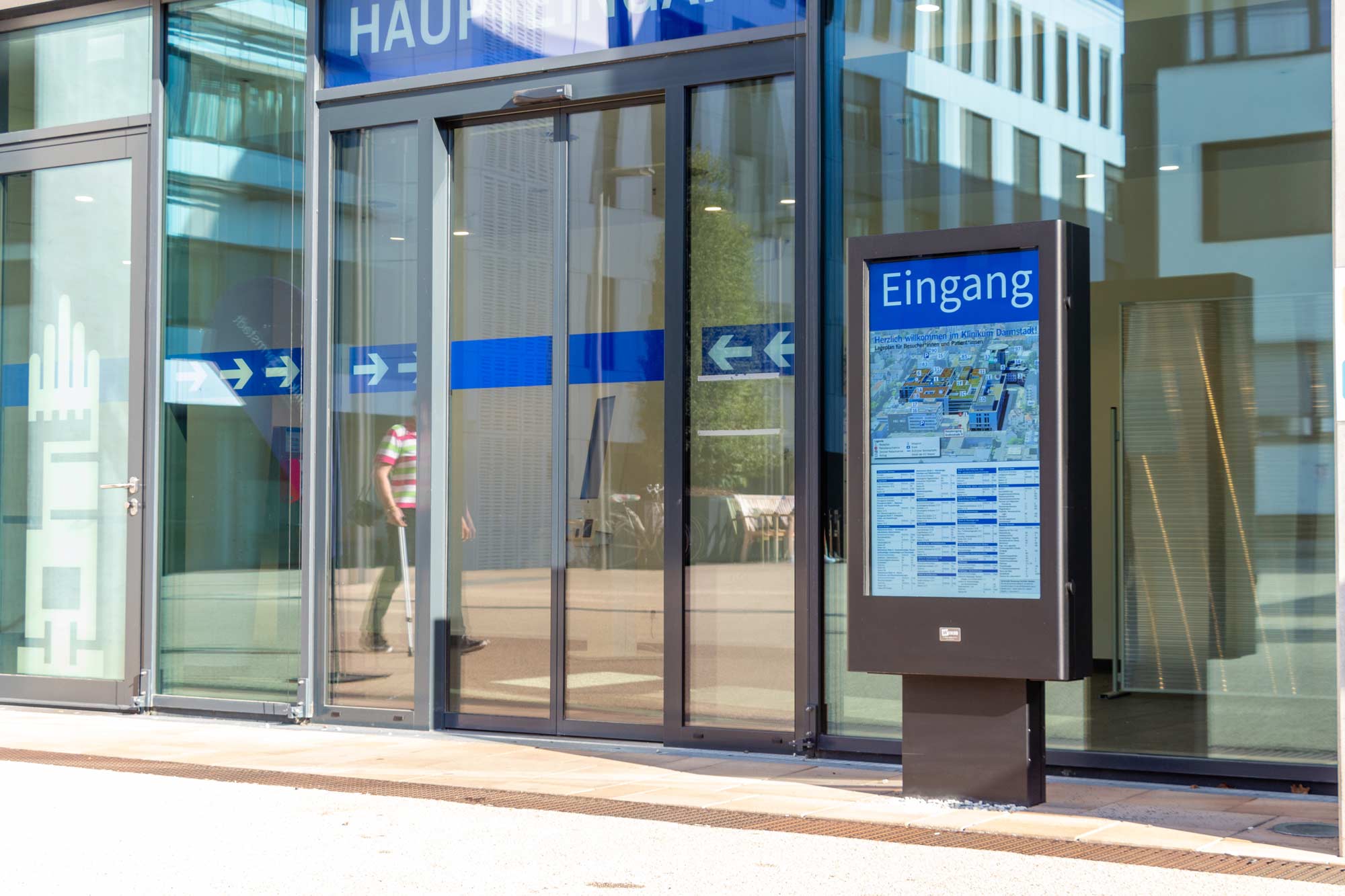 Eingang mit Glastüren eines modernen Gebäudes, daneben ein Informationsdisplay mit blauem Schild und Gebäudeplan.