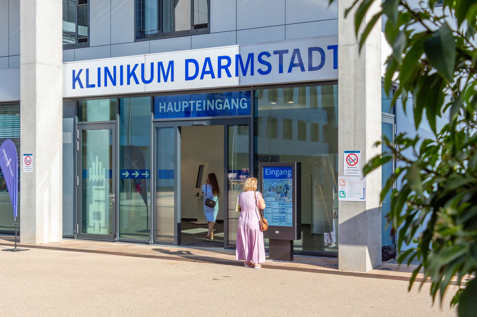 Haupteingang des Klinikums Darmstadt mit zwei Frauen, eine liest am Infostand und eine geht durch die Glastür.