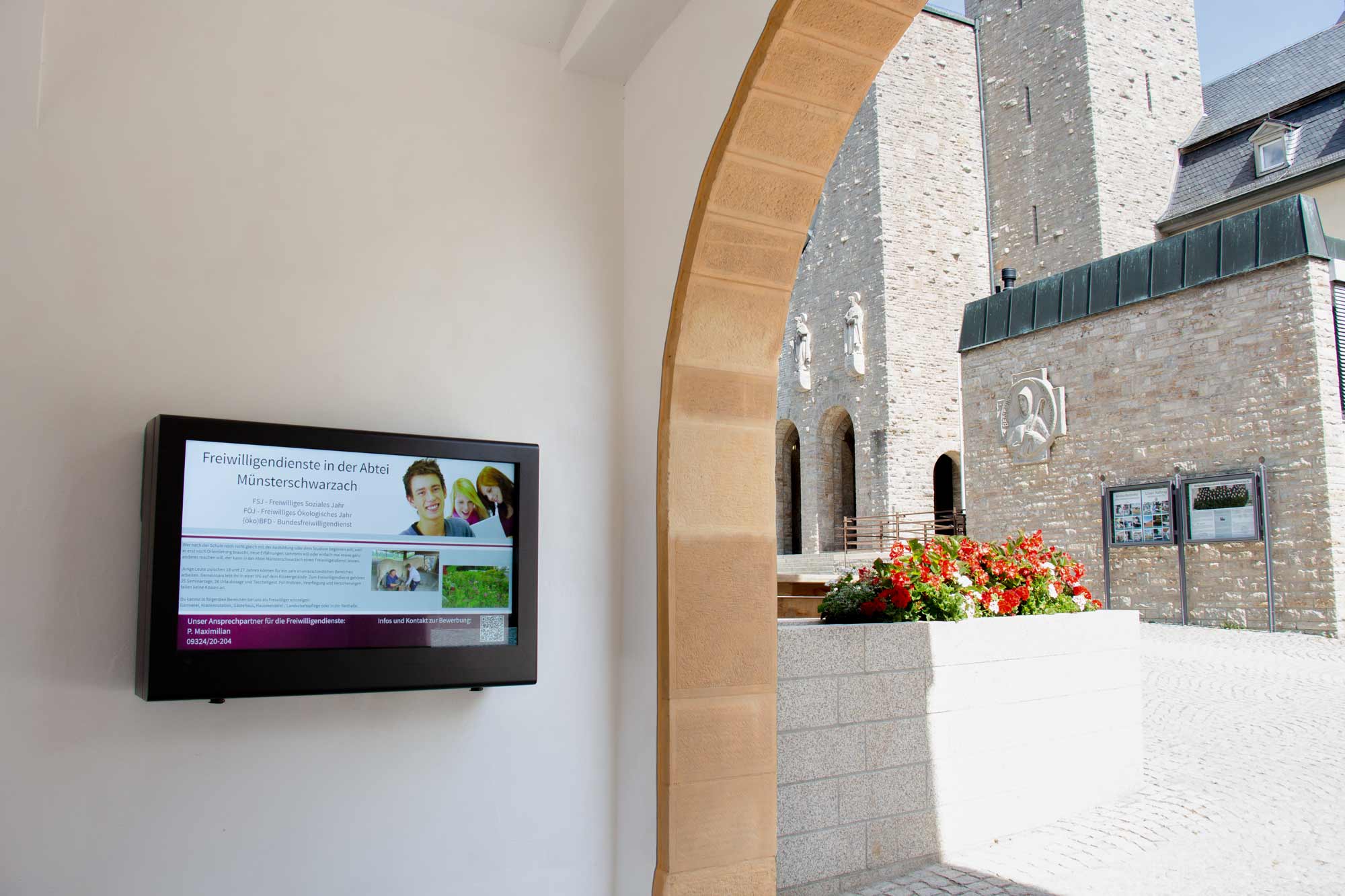 Digitale Informationsanzeige zur Freiwilligendienst im Kloster Münsterschwarzach an weißer Wand neben Sandsteinbogen mit Blick auf Innenhof und Blumen.