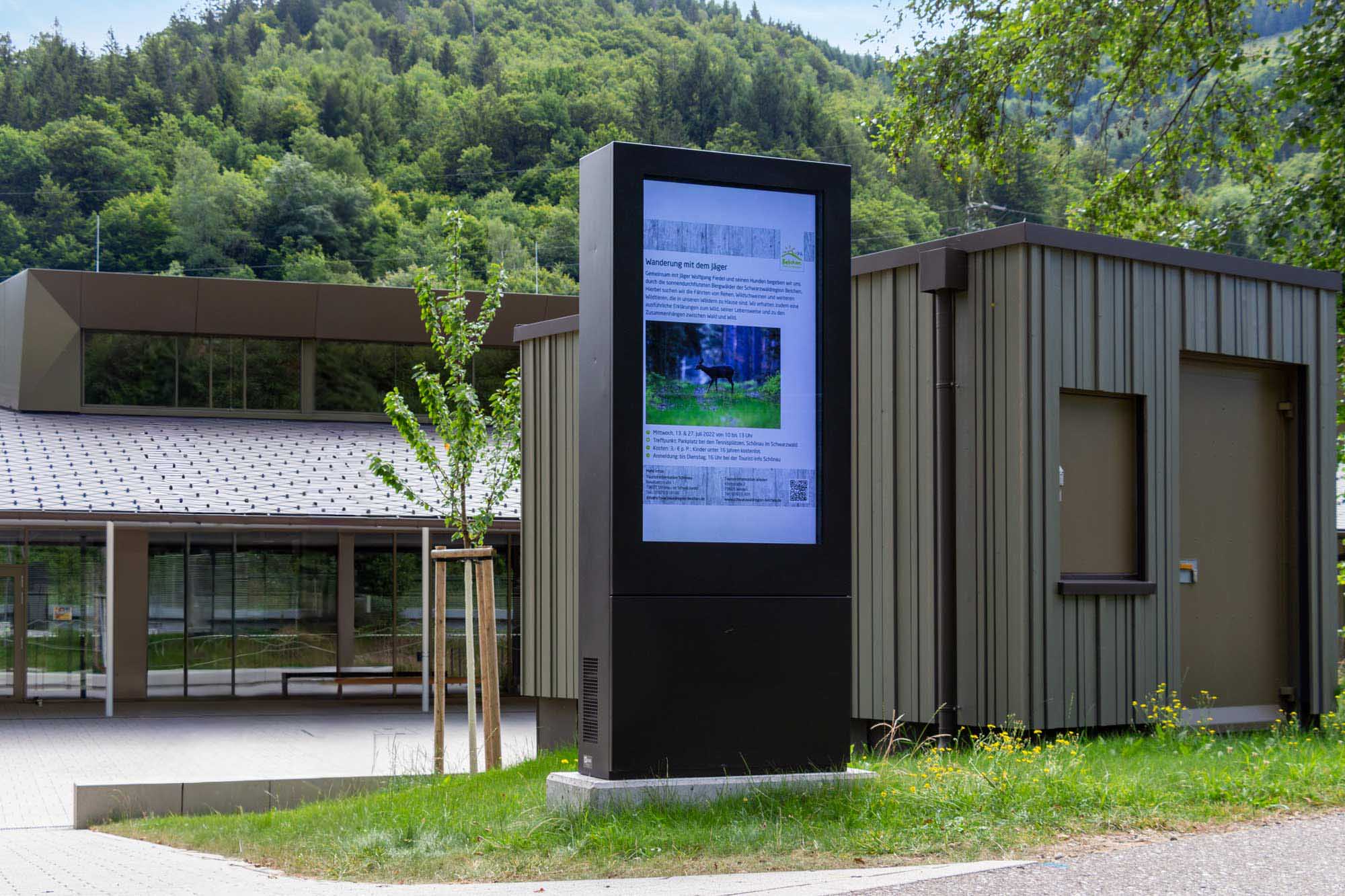 Digitale Informationsstele mit Ankündigung eines Wanderungsevents vor modernem Gebäude und bewaldetem Hügel im Schwarzwald.