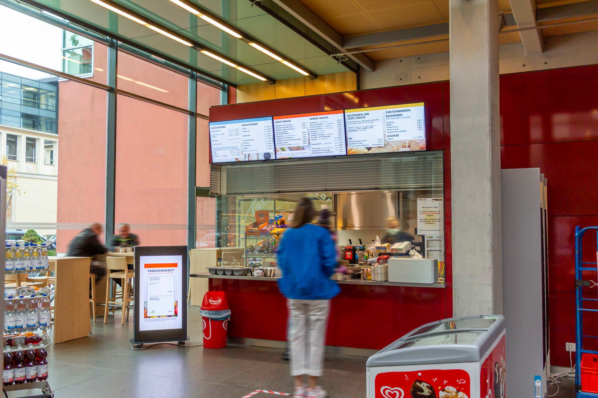 Menschen stehen vor einem Verkaufsfenster mit digitaler Speisekarte und Kühlschrank mit Eis in moderner Cafeteria.
