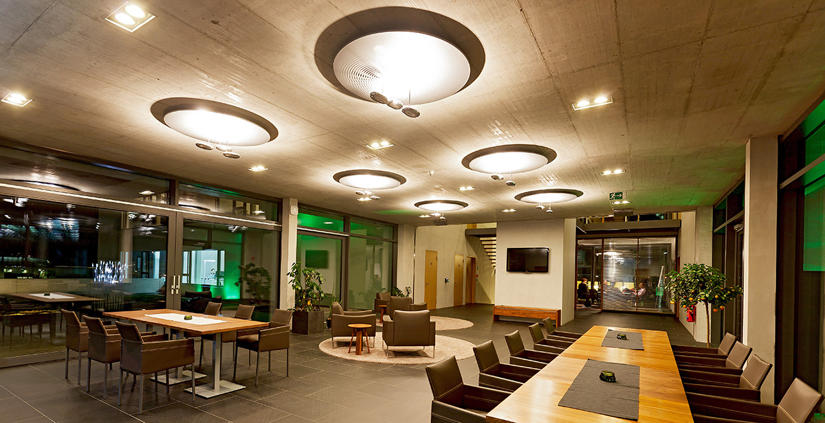 Moderner Lounge-Bereich mit großen Holztischen, bequemen Stühlen, Pflanzen und runden Deckenleuchten in einem Raum mit großen Fenstern.