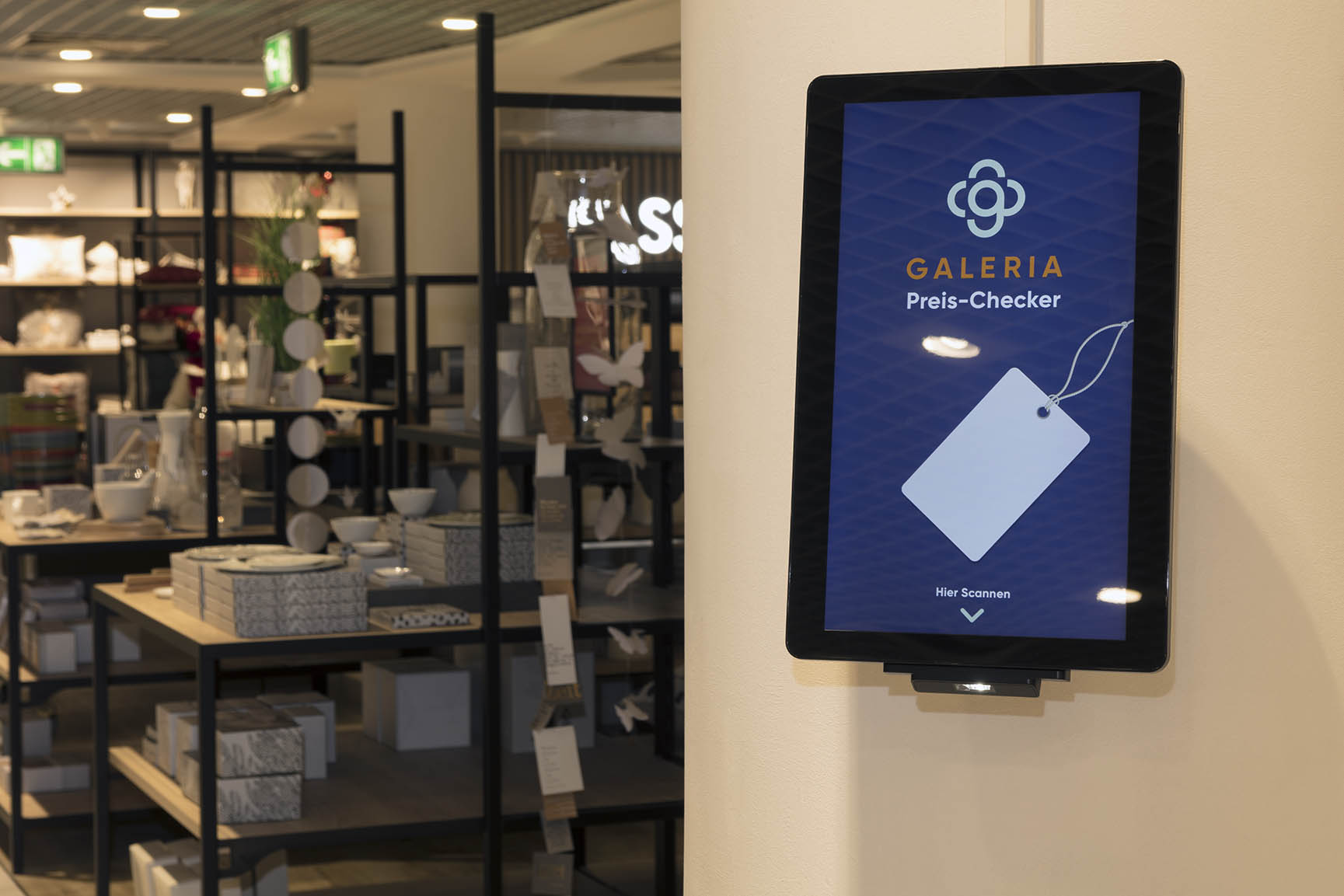 Digitaler GALERIA Preis-Checker Bildschirm an einer Wand in einem Geschäft mit Regalen und Haushaltsartikeln im Hintergrund.