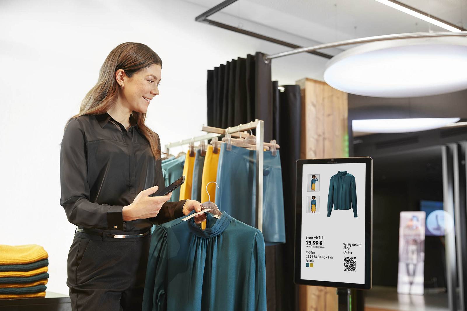 Frau scannt mit ihrem Smartphone eine grüne Bluse, präsentiert auf einem Kleiderbügel im Geschäft, neben einem digitalen Info-Display mit Produktdetails.