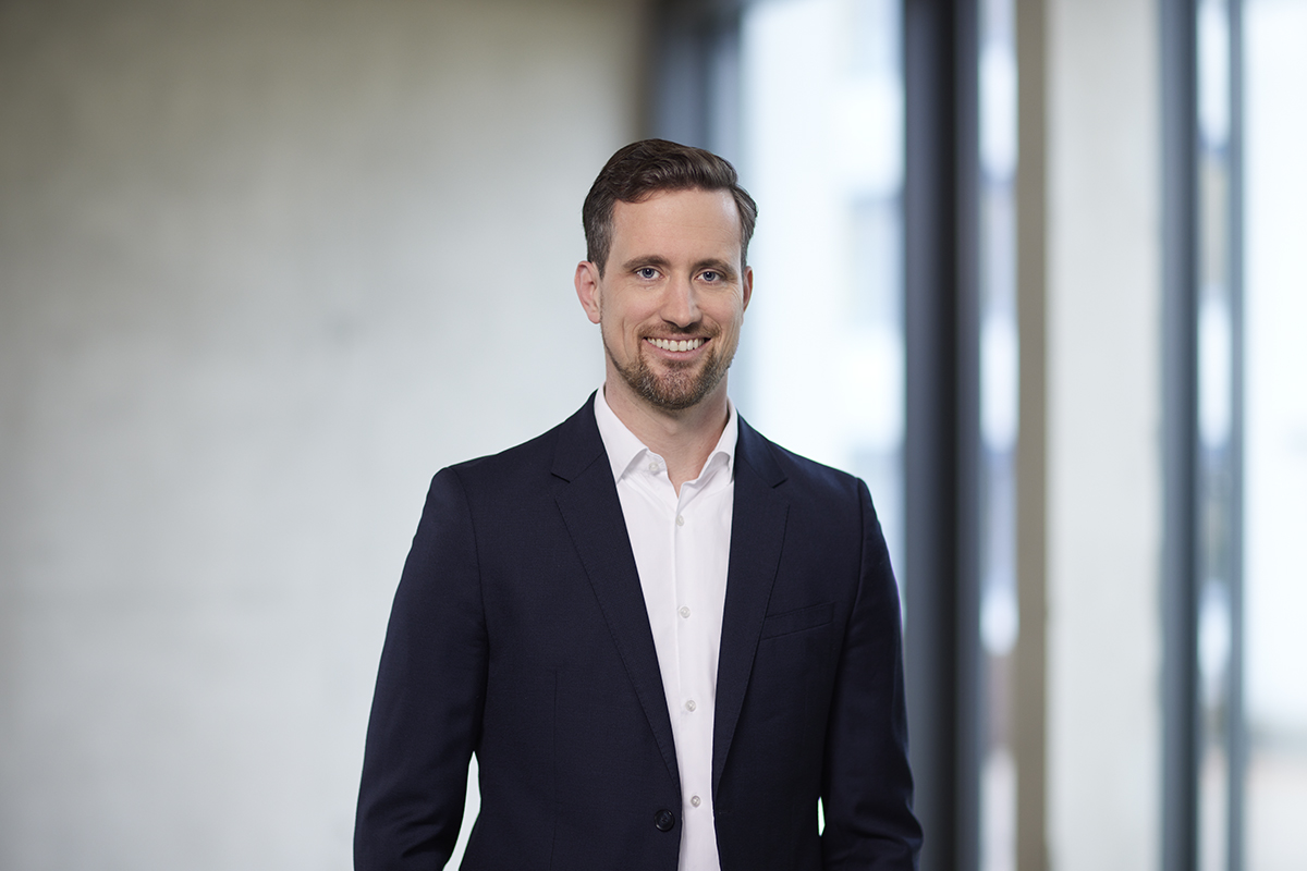 Lächelnder Mann in dunkelblauem Anzug und weißem Hemd vor unscharfem, modernem Fensterhintergrund.