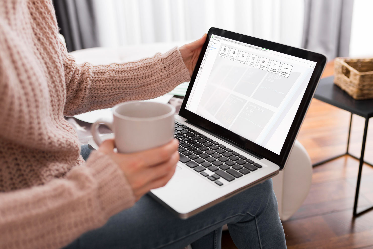 Person sitzt auf einem Stuhl, hält eine Tasse und benutzt einen Laptop mit geöffneter Software-Oberfläche.