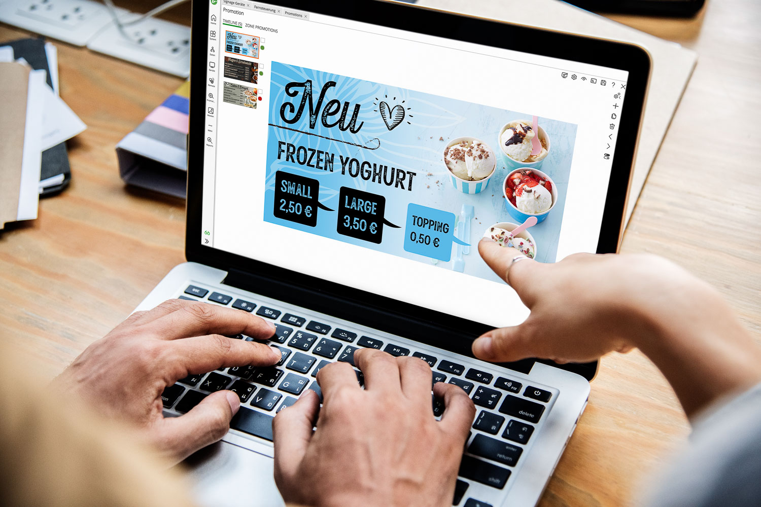 Zwei Personen arbeiten an einem Laptop, das auf dem Bildschirm ein Werbedesign für gefrorenen Joghurt mit Preisen anzeigt.