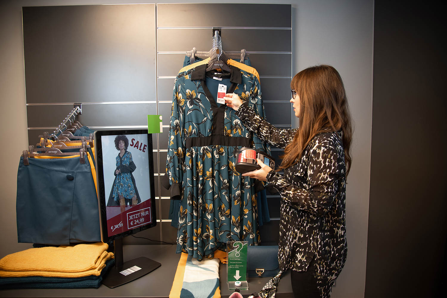 Frau in einem Geschäft zeigt ein Preisschild an einem geblümten Kleid neben einem digitalen Verkaufsdisplay mit Rabattangeboten.