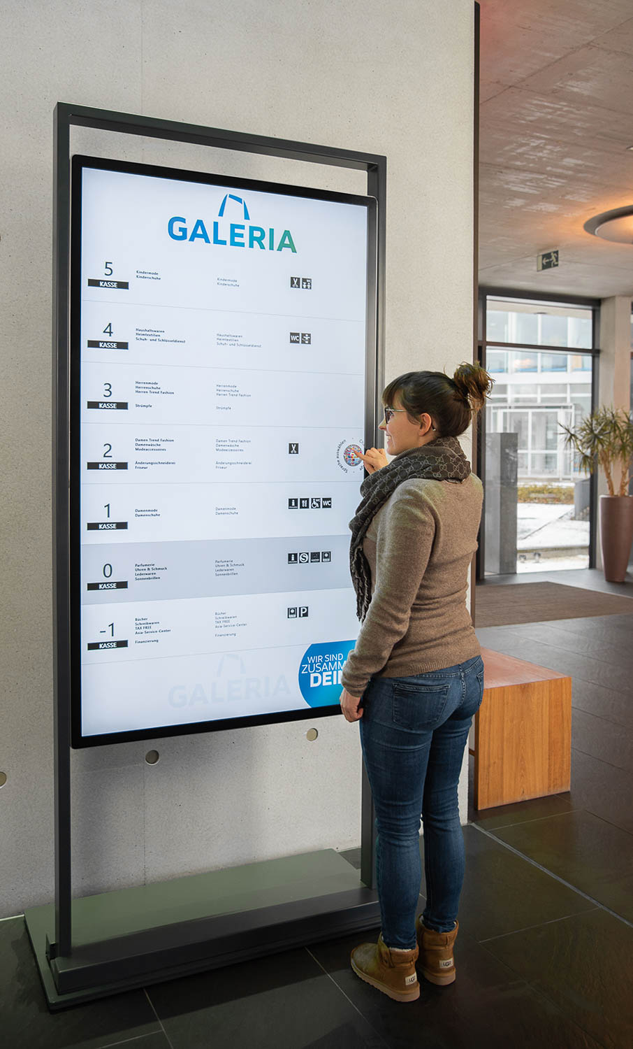 Frau liest an einem digitalen Verzeichnis im Eingang eines modernen Gebäudes die Stockwerkübersicht und Abteilungen von GALERIA.