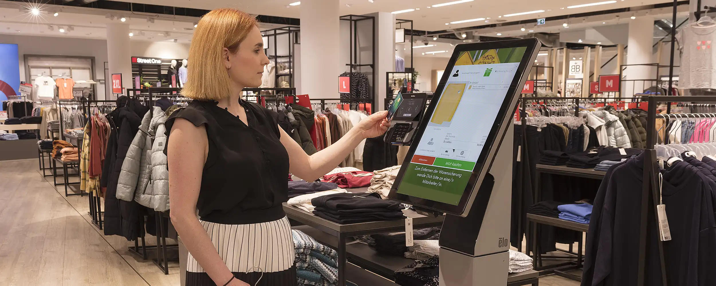 Frau scannt Kleidung und zahlt mit Karte an einem Self-Checkout-Terminal in einem Bekleidungsgeschäft.