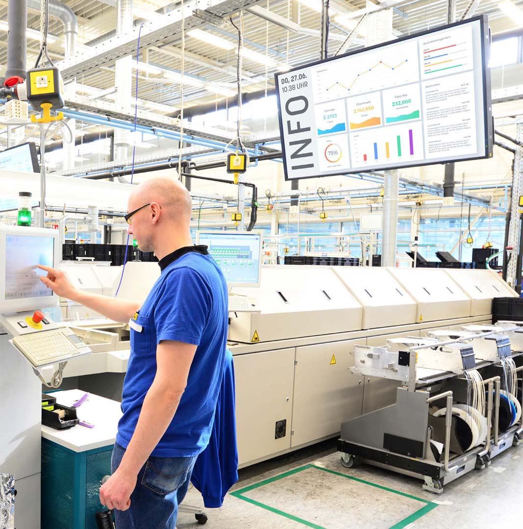 Mann in blauem T-Shirt bedient einen Touchscreen in einer hellen Produktionshalle mit großen Maschinen und Informationsbildschirmen.