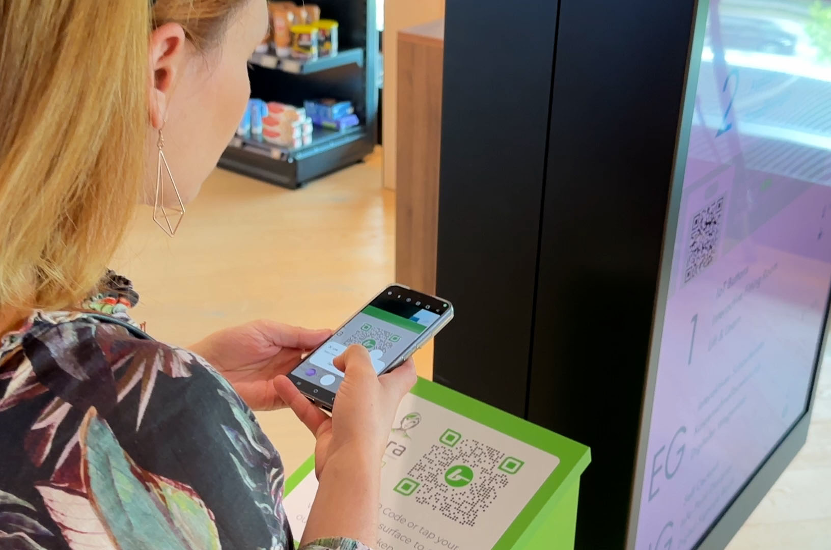 Frau scannt mit Smartphone einen auf grüner Fläche gedruckten QR-Code in einem Innenraum.