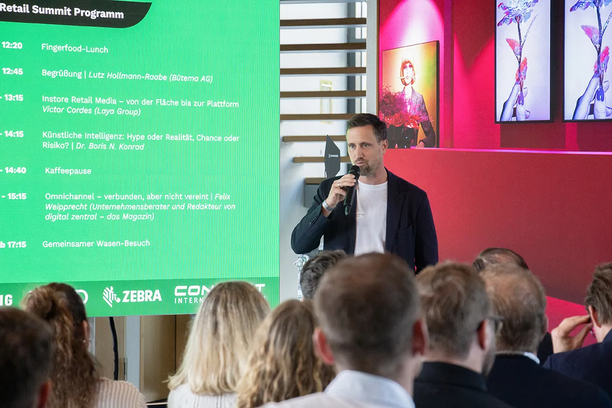 Mann hält Mikrofon und spricht vor Publikum neben einer grünen Präsentationstafel mit Programm des Retail Summit.