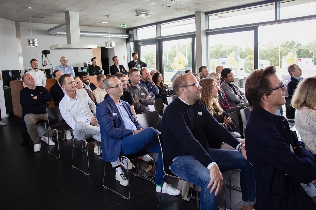 Eine Gruppe von Menschen sitzt aufmerksam in einem modernen Veranstaltungsraum mit großen Fenstern.