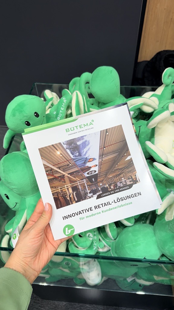 Hand hält eine Broschüre von BÜTEMA über innovative Retail-Lösungen vor einem Glasbehälter mit grünen Stoffpuppen.